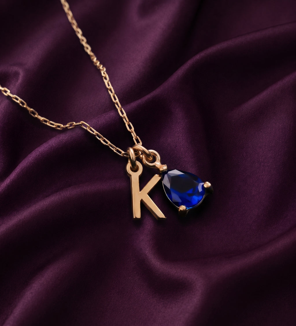 Necklace with a K initial charm and a blue pear-shaped stone on purple satin